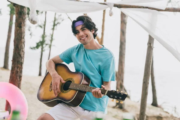 Portrait of attractive cheerful guy playing guitar relax enjoying good weather sunlight abroad at beach picnic outdoors.
