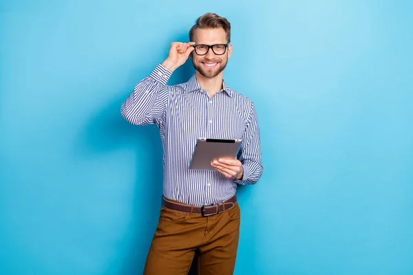 Portrait of positive friendly man hand touch glasses hold tablet look camera isolated on blue color background.
