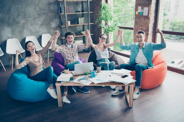 Full size photo of overjoyed delighted people sitting beanbag hold hands raise fists celebrate achievement indoors.