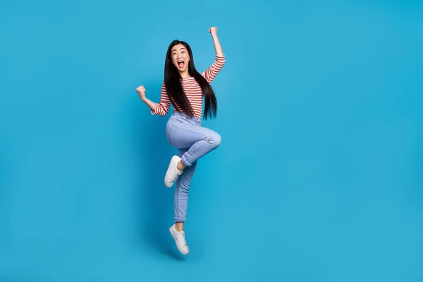 興奮した幸運な女性の完全な長さの写真は 高い上昇拳をジャンプストライプシャツを身に着けています空のスペース隔離された青の背景 — ストック写真