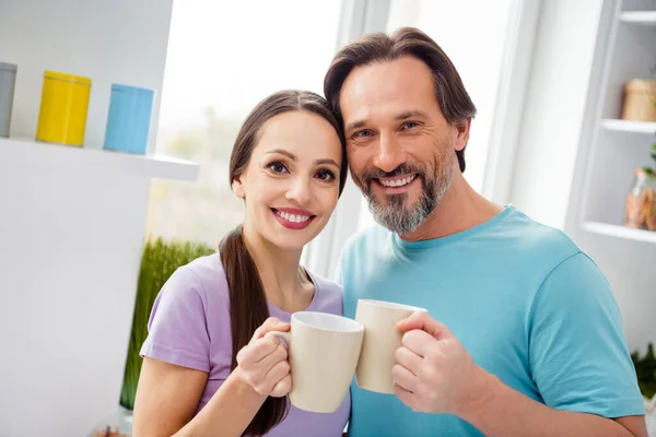 Portrait of positive cheerful people hug touch heads hold clink coffee cup toothy smile kitchen indoors.
