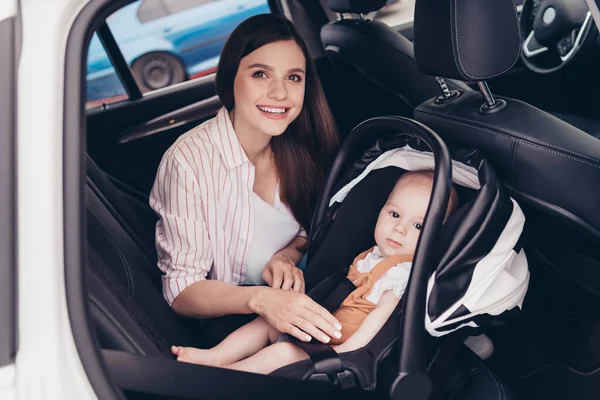 Photo of positive charming mother adorable baby child sitting safety chair automobile salon prepare travel.