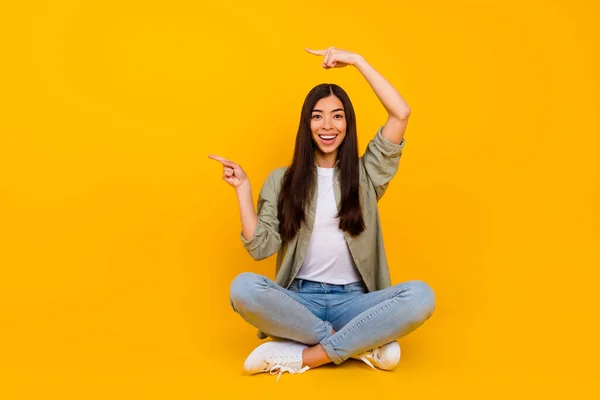 Full body portrait of excited funny girl sit floor indicate fingers empty space blank isolated on yellow color background.