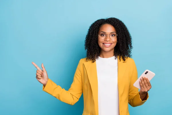 Photo of young black woman happy positive smile hold mobile point finger empty space ad promo isolated over blue color background.