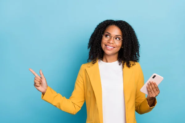 Photo of young black woman happy positive smile indicate finger empty space promo choice cellphone isolated over blue color background.