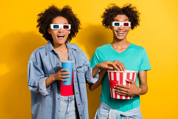 Photo of amazed afro american people couple wear 3d glasses fly pop corn drink isolated on yellow color background.