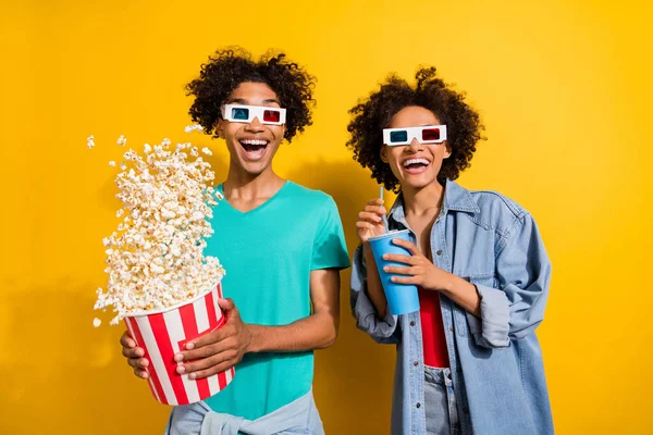 Photo of amazed happy dark skin people couple film glasses pop corn fly drink isolated on yellow color background.