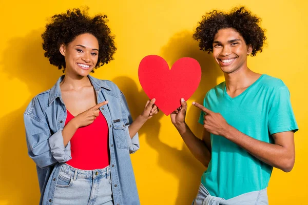 Photo of happy dark skin man and woman people couple point finger heart isolated on yellow color background.
