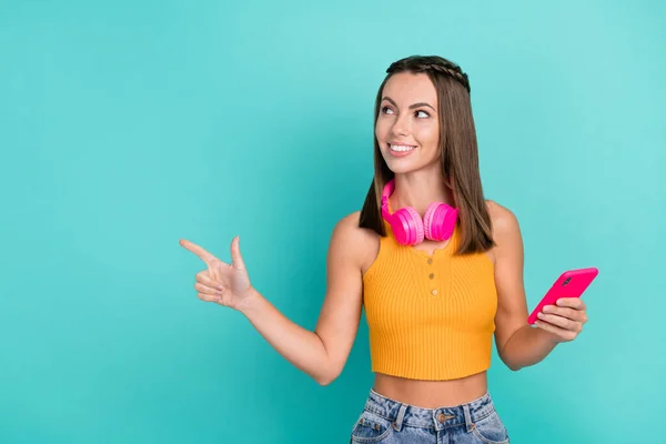 Photo of happy brunette trendy stylish woman point finger empty space look sale hold phone wireless headset isolated on teal color background.