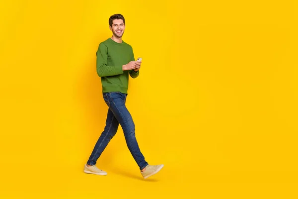 Photo of charming pretty guy dressed green pullover writing modern device walking empty space isolated yellow color background.