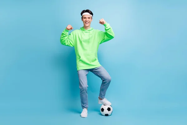 Photo of cool confident young guy dressed green hoodie playing football showing strength smiling isolated blue color background.