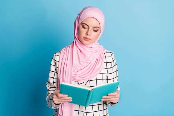 Photo of young attractive muslim woman serious read book story novel wear pink hijab isolated over blue color background.