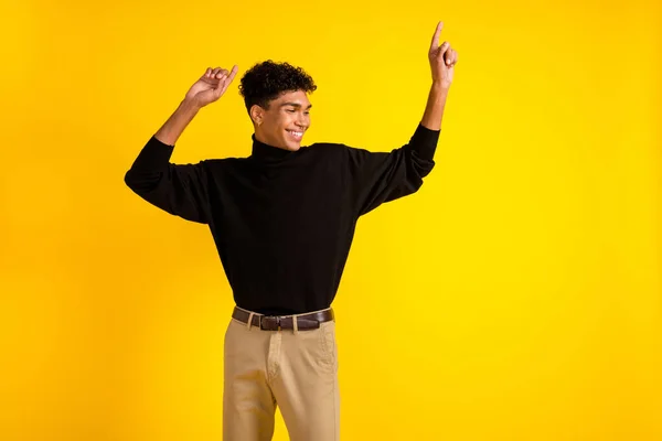 Photo of funky charming dark skin man wear black sweater dancing pointing empty space isolated yellow color background.