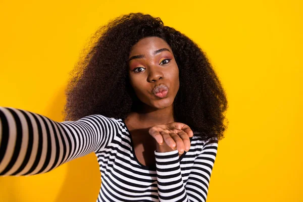 Photo portrait of curly woman taking selfie wearing stylish clothes sending air kiss isolated vibrant yellow color background.