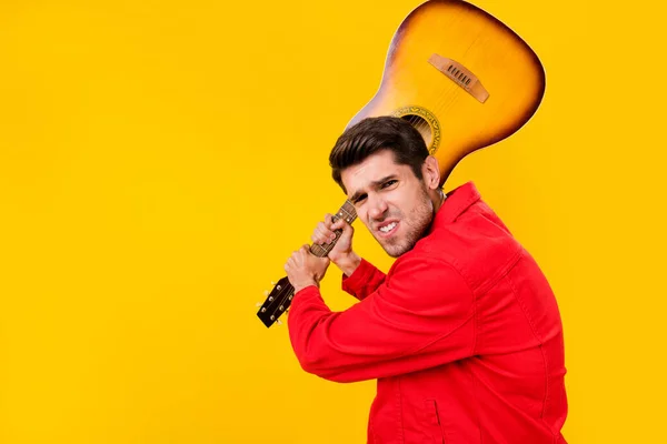 Portrait of attractive rude mad guy holding guitar fighting isolated over bright yellow color background.