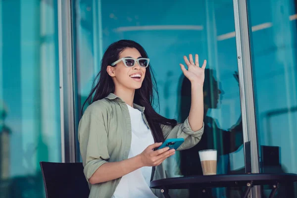 Photo of sweet excited lady dressed casual clothes dark eyewear sitting cafeteria waving arm modern gadget outside urban town house.