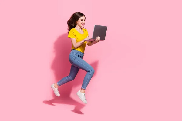 Full body profile photo of sweet young brunette lady run with laptop wear t-shirt jeans shoes isolated on pink background.