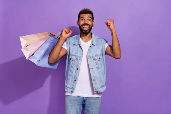 Photo of funny lucky man wear jeans waistcoat holding shoppers rising fist isolated violet color background.