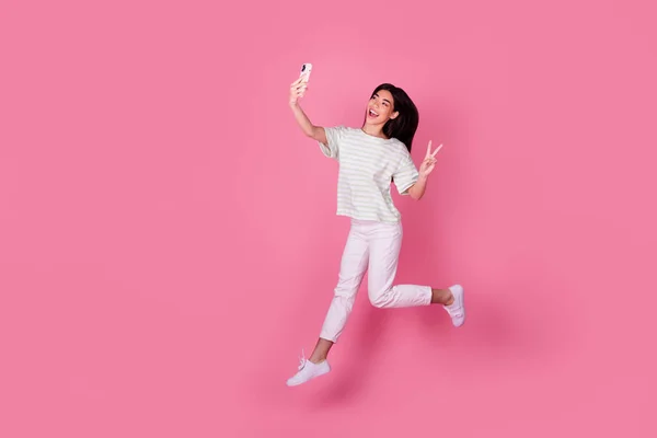 Photo of cute young japanese lady hold gadget take selfie jump show v-sign dressed stylish white clothes isolated on pink color background.