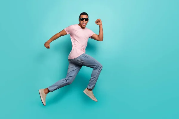 Full length photo of excited pretty guy wear pink t-shirt jumping high running empty space isolated teal color background.