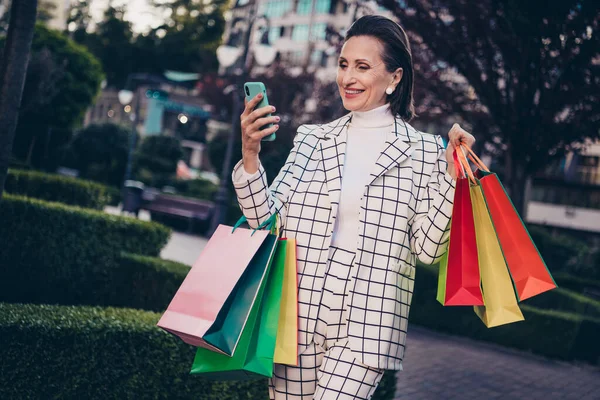 Photo of cute sweet woman wear plaid jacket typing modern device holding shoppers outside city residential complex.