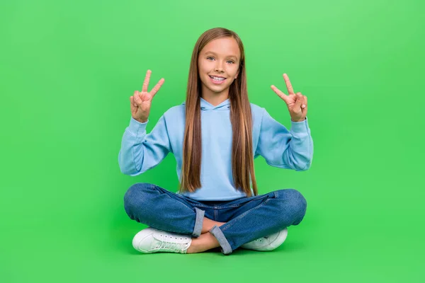 Full size photo of lovely small girl sit floor legs crossed double v-sign dressed trendy blue sweatshirt isolated on green color background.