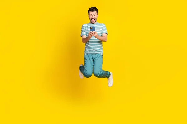 Full size photo of ecstatic impressed guy striped t-shirt blue pants sneakers look on phone jumping isolated on yellow color background.