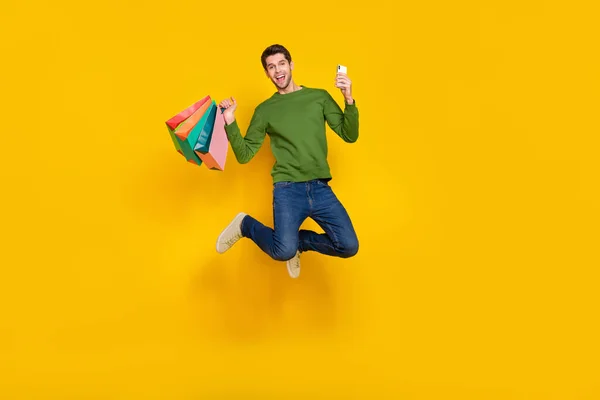 Photo of impressed pretty guy wear green sweater jumping rising shoppers chatting gadget isolated yellow color background.