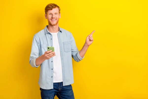 Photo portrait of handsome young man hold smart phone point empty space dressed trendy denim garment isolated on yellow color background.