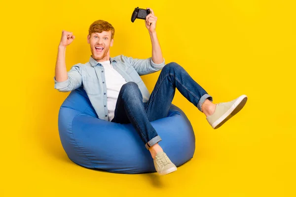 Photo of lucky funny man wear jeans shirt sitting beanbag rising fists playstation empty space isolated yellow color background.