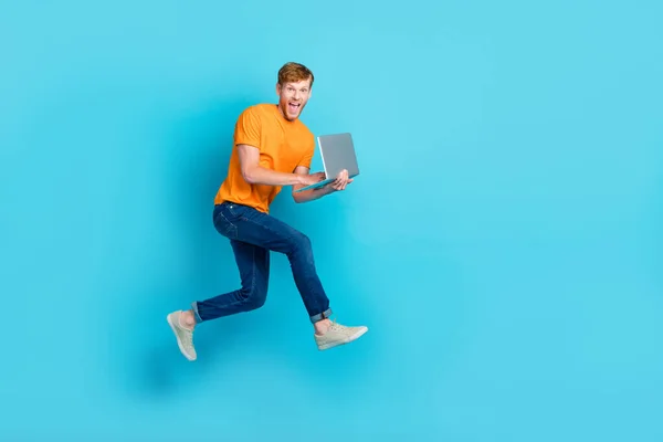 Full size profile photo of hooray red hair guy run with laptop wear t-shirt jeans boots isolated on blue color background.