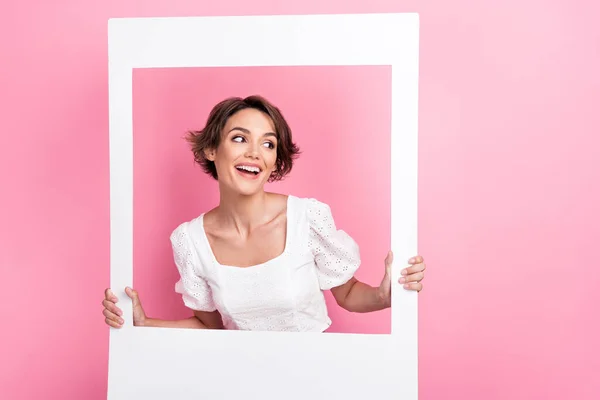 Photo of charming excited lady wear white blouse looking inside photo frame empty space isolated pink color background.