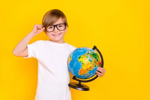 Portrait of handsome cheerful diligent pre-teen boy holding globe touching specs isolated over bright yellow color background.