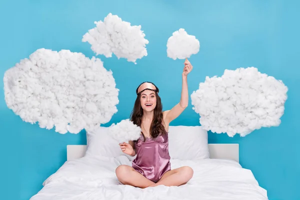 Photo of shiny cute woman dressed pink pajama sitting bed holding clouds isolated blue color background.