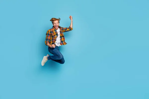 Photo of astonished cheerful guy with red hair dressed checkered shirt jumping raise fist up scream yeah isolated on blue color background.
