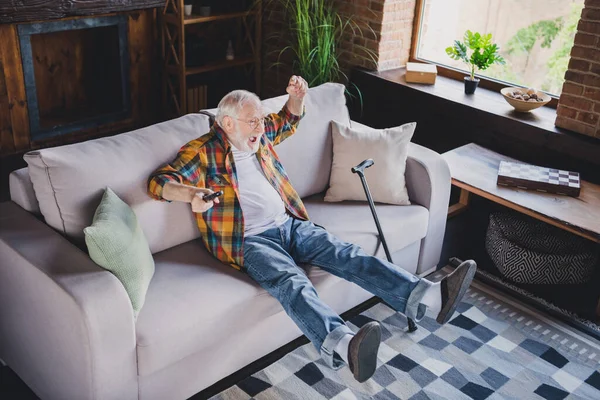 Photo of funny lucky retired man dressed plaid shirt watching soccer sitting couch holding nursery cane indoors apartment.
