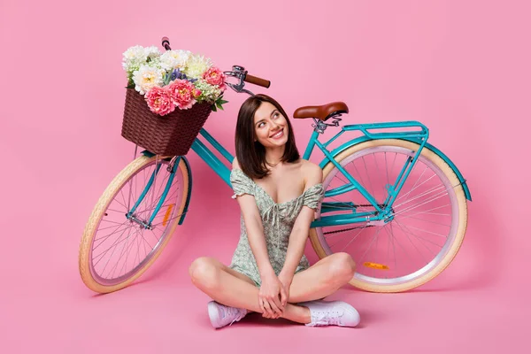 Full body photo of cute lady sit near bicycle look up wear printed dress boots isolated on pink color background.