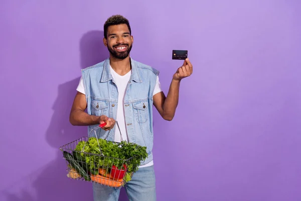 Photo of cute pretty guy dressed denim vest holding green veggies basket credit card empty space isolated purple color background.