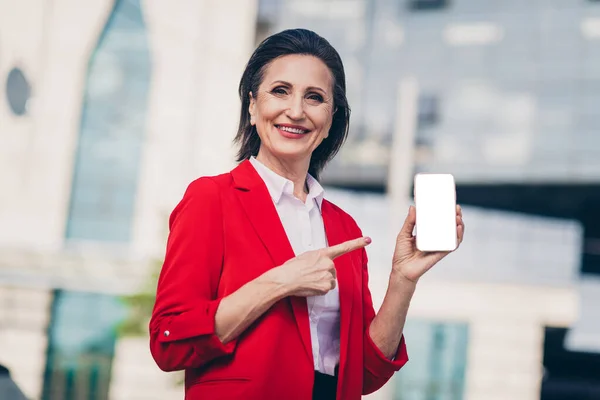 Photo of good mood pretty senior manager woman dressed red blazer pointing modern device empty space outdoors urban town street.