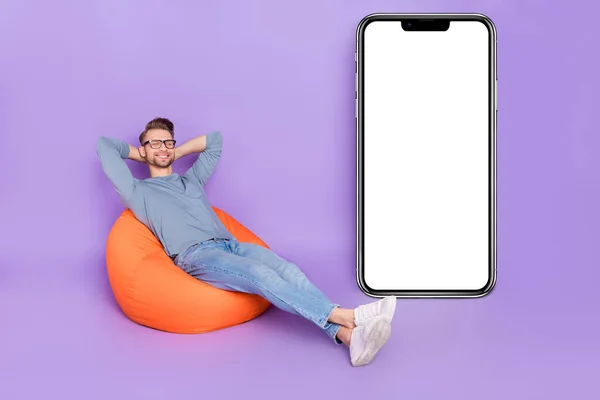 Photo of dreamy pretty guy dressed grey shirt sitting bean bag arms behind head gadget empty space isolated purple color background.