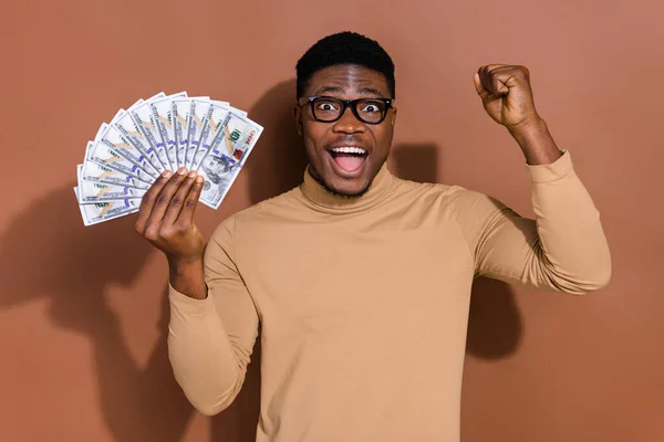 Portrait of attractive cheerful lucky guy holding fan cash atm us dollars having fun rejoicing isolated over brown color background.