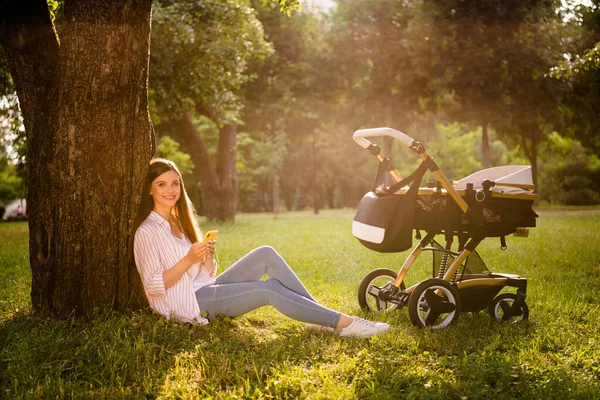 Full length photo of cute mom sit lawn urban street park use gadget blog while infant kid sleep pram outdoors.