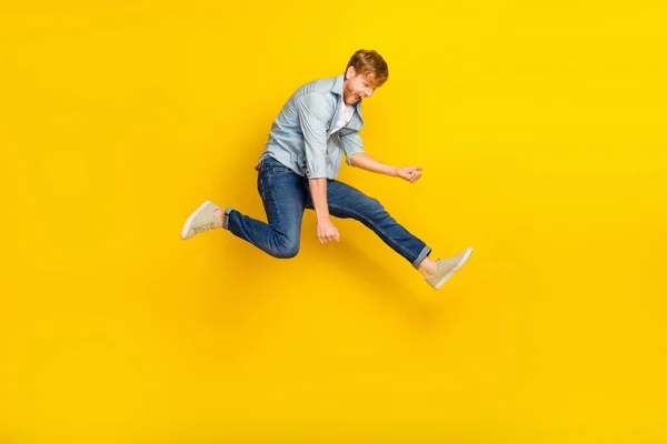 Full size photo of handsome guy ginger hairdo blue shirt jeans sneakers jump play imaginary guitar isolated on yellow color background.