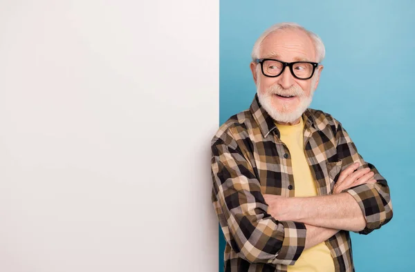 Photo of funky pretty man pensioner dressed checkered shirt eyewear arms folded billboard empty space isolated blue color background.