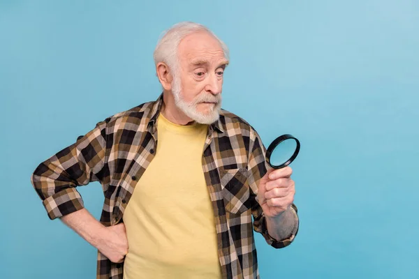 Photo of funky interested man pensioner dressed checkered shirt looking enlarging glass empty space isolated blue color background.