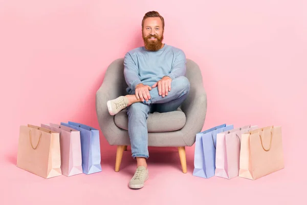 明るい男座っている椅子の残りの部分の完全な長さの肖像ピンクの色の背景に隔離されたショッピングパッケージをリラックス — ストック写真