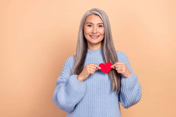 Photo of charming good mood mature lady hold little red paper heart take care of her health isolated on beige color background.