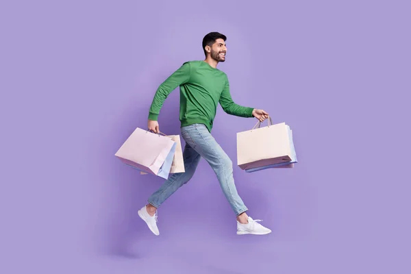 年轻男子的全身照片 提着购物袋匆忙销售 穿着时髦的绿色衣服 背景为紫罗兰色 — 图库照片