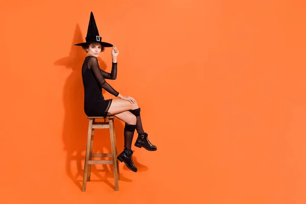 フルボディ写真の素敵な若い女の子浮気見るタッチ帽子スツール着用スタイリッシュな黒ハロウィン魔女見て隔離されたオレンジ色の背景 — ストック写真