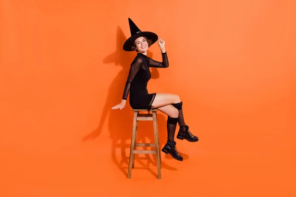 魅力的な女性の完全な長さの写真あなたの笑顔座ってバーチェアはオレンジの色の背景に隔離された流行の黒ハロウィンの服を着て — ストック写真